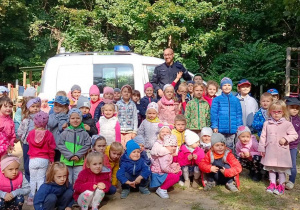 Dzieci wraz z Panem Policjantem pozują na tle radiowozu w ogrodzie przedszkolnym.