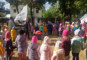 Dzieci zebrane w kole w ogrodzie przedszkolnym. Pośrdoku na ławce stoi odnaleziona wcześniej skrzynia ze skarbem. Nauczycielki trzymają w rękach pirackie flagi.