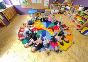 Na podłodze w sali rozłożona jest chusta animacyjna, dzieci leżą na niej z podniesionymi nogami.