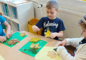 Dzieci siedzą przy stolikach. Na kolorowych kartkach przyklejają różne liście.