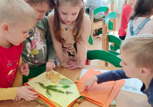 Dzieci stoją wokół stolika. Na stoliku leży zielnik, dzieci oglądają jego strony.