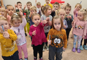 Zdjęcie zbiorowe. Dzieci stoją w sali każde trzyma kredkę przed sobą w wyciągniętej do przodu ręce.