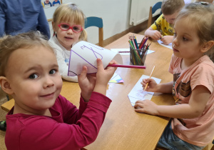 Trzy dziewczynki siedzą przy stoliku. Kolorują sylwety kredek ze swoimi imionami. Środkowa dziewczynka patrzy w obiektyw i prezentuje swoją pracę.