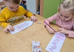 Dzieci siedzą przy stoliku patrzą i kolorują na sylwety kredek ze swoimi imionami.