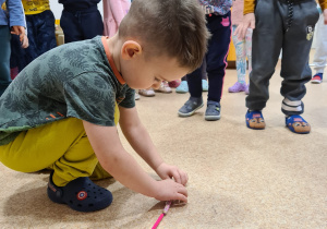 Chłopiec układa na podłodze kredki jedna za drugą. Reszta dzieci stroi za chłopcem przyglądają się czy polecenie zostało wykonane poprawnie.
