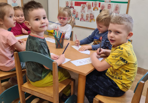 Dzieci siedzą przy stoliku patrzą i kolorują na sylwety kredek ze swoimi imionami.