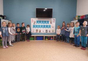 Grupa dzieci stoi przy tablicy, na której widnieje napis i zawieszone są ilustracje z PRAWAMI DZIECKA.
