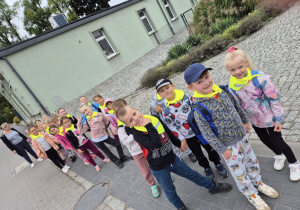Dzieci w odblaskowych apaszkach stoją w parach przed przedszkolem.