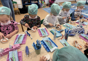 Dzieci w czepkach i maskach bawią się narzędziami stomatologicznymi.