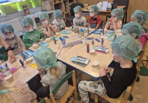 Dzieci w czepkach i maskach bawią się narzędziami stomatologicznymi.