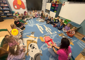 Dzieci siedzą w kręgu na dywanie, słuchają wykładu prowadzącego.