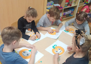 Dzieci tworzą pracę plastyczną, wyklejają papierem dynie przy stolikach.