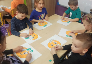 Dzieci tworzą pracę plastyczną, wyklejają papierem dynie przy stolikach.