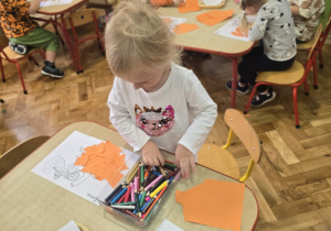 Dzieci tworzą pracę plastyczną, wyklejają papierem dynie przy stolikach.