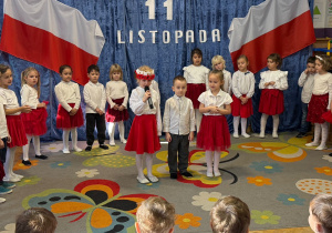 Zdjęcie przedstawia dzieci w biało-czerwonych strojach. Stoją w półkolu, a trzy osoby na środku.
