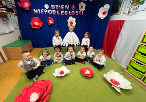 Na zdjęciu grupa maluszków, Część dzieci siedzi na dywanie część stoi za nimi. Jedno dziecko jest przebrane za orła w złotej koronie.