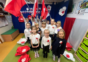 Zdjęcie grupowe starszaków. Dzieci stoją na tle dekoracji z okazji Święta Niepodległości. Dzieci stojące z tyłu trzymają duże flagi.