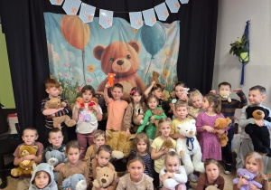 Dzieci pozują do zdjęcia grupowego na tle dużej ilustracji z misiem