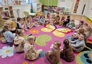 dzieci siedzą na kolorowym dywanie w kole, słuchają nauczyciel
