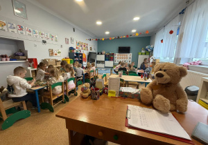 zdjęcie przedstawia obraz całej sali, na pierwszym planie jest biurko nauczyciela na którym leży miś. Dzieci siedzą przy stolikach