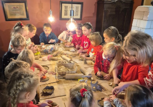 Dzieci siedzą przy długim stole, na którym leżą wałki i glina