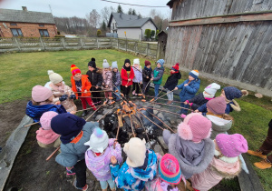 dzieci w dużym kole stoją wokół ogniska i pieką kiełbaski na patykach