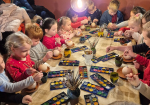 dzieci siedzą przy długim stole i malują farbami dzieła z gliny