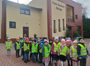 Dzieci w kamizelkach odblaskowych stoją na tle budynku Domu Seniora "Akacja"