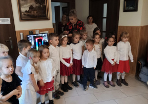 Dzieci w galowych strojach stoją w półkolu świetlicy Domu Akacja, śpiewają piosenki patriotyczne