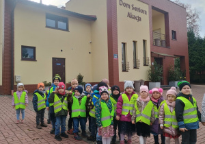 Dzieci w kamizelkach odblaskowych stoją na tle Domu Seniora Akacja