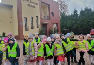 Dzieci w kamizelkach odblaskowych stoją na tle Domu Seniora Akacja