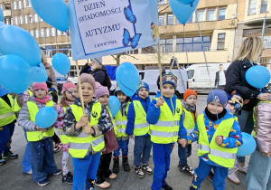 Dzieci stoją na Pasażu Schillera w Łodzi ubrane w żółte kamizelki trzymają niebieskie balony z helem i transparenty które głoszą hasła zrozumienia dla dzieci w spektrum autyzmu.