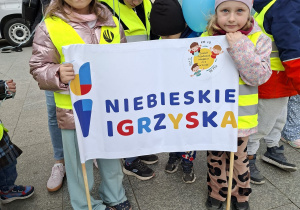Dzieci stoją na Pasażu Schillera w Łodzi ubrane w żółte kamizelki trzymają niebieskie balony z helem i transparent z napisem "Niebieskie Igrzyska"
