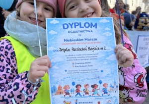 dwie dziewczynki trzymające niebieskie balony pozują do zdjęcia trzymając dyplom uczestnictwa w Niebieskim Marszu