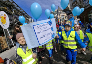 Dzieci stoją na Pasażu Schillera w Łodzi ubrane w żółte kamizelki trzymają niebieskie balony z helem i transparenty które głoszą hasła zrozumienia dla dzieci w spektrum autyzmu.