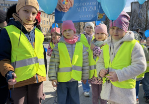 Dzieci stoją na Placu Wolności w Łodzi ubrane w żółte kamizelki trzymają niebieskie balony z helem i transparenty które głoszą hasła zrozumienia dla dzieci w spektrum autyzmu.